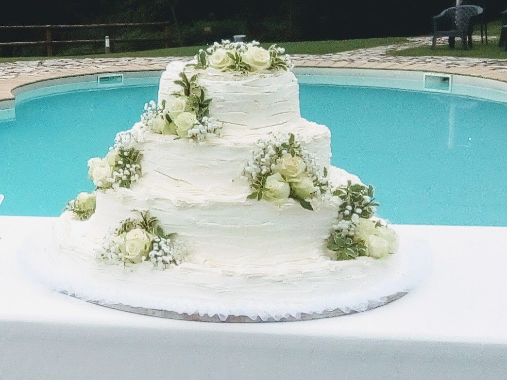 Torta nuziale a tre piani decorata con fiori bianchi, situata a bordo piscina.