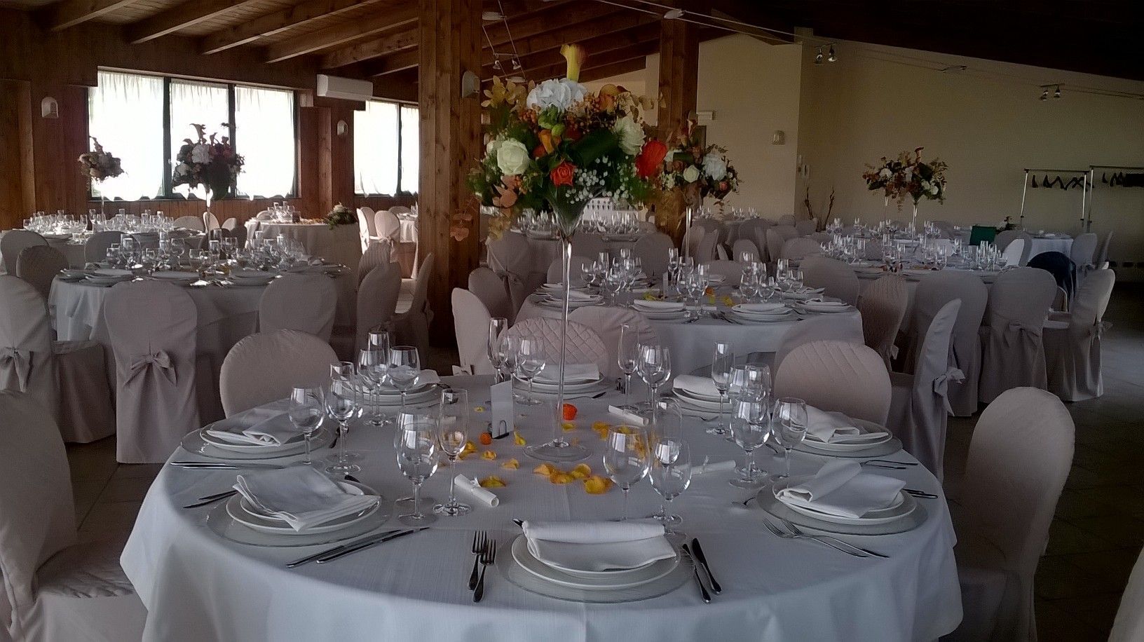 Elegante sala per banchetti con tavoli rotondi apparecchiati per un evento formale. Tavoli bianchi, sedie con coprisedie e grandi composizioni floreali.