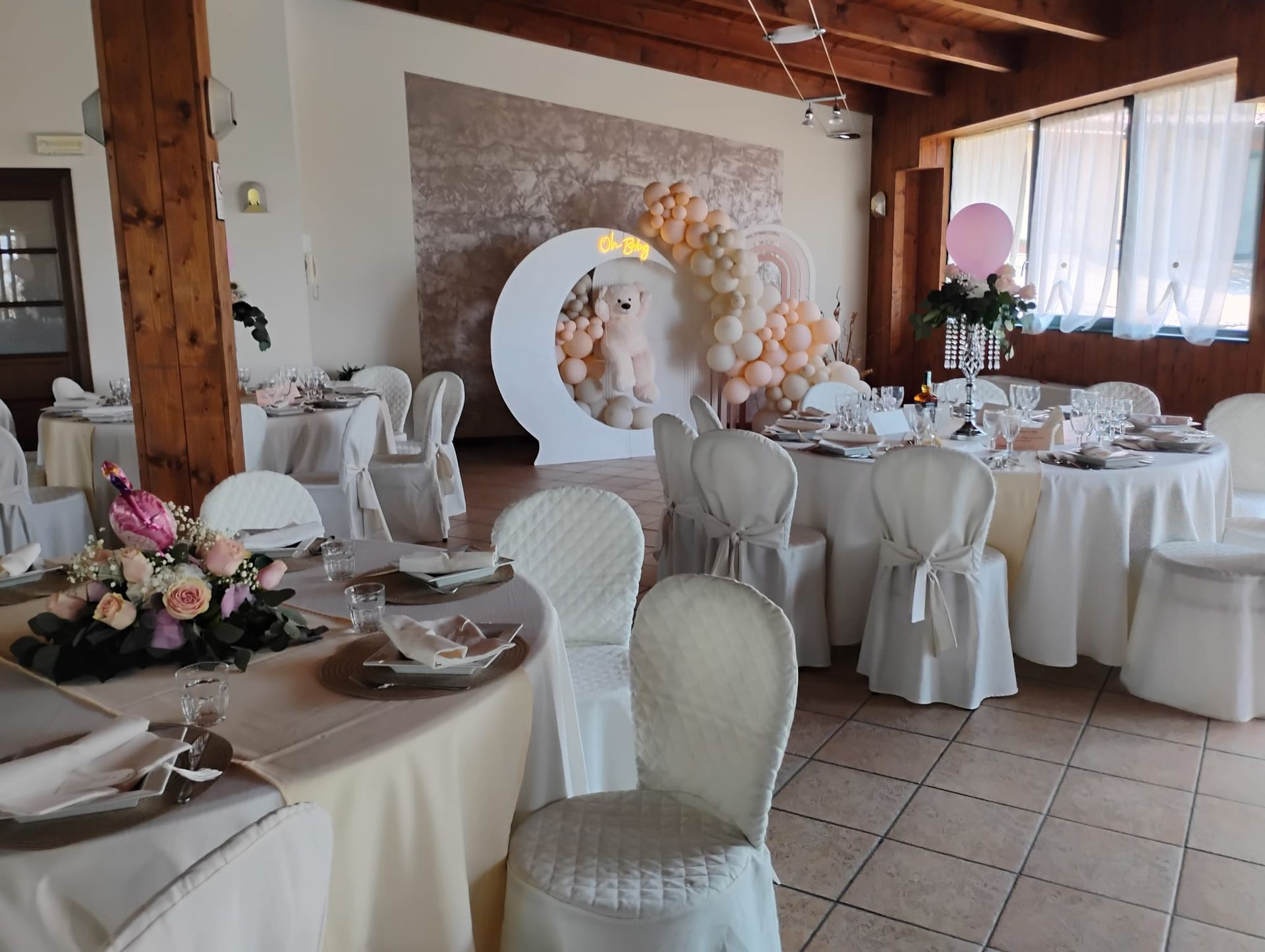 Sala ricevimenti decorata per una festa: tavoli rotondi con tovaglie bianche, sedie con coprisedie, arco di palloncini.