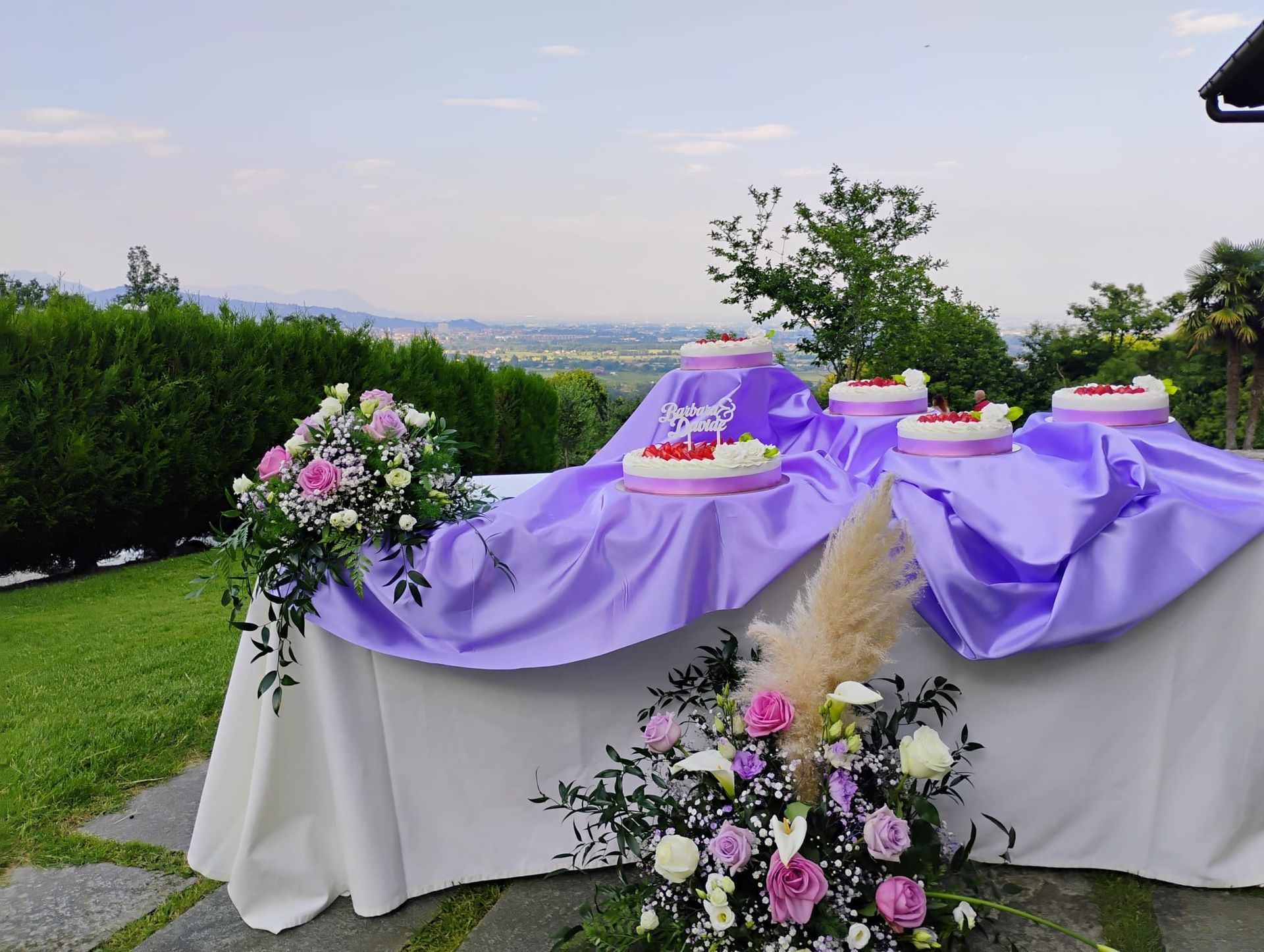 Tavolo per la torta nuziale con tovaglia viola, torta bianca, composizioni floreali e vista sulle montagne.