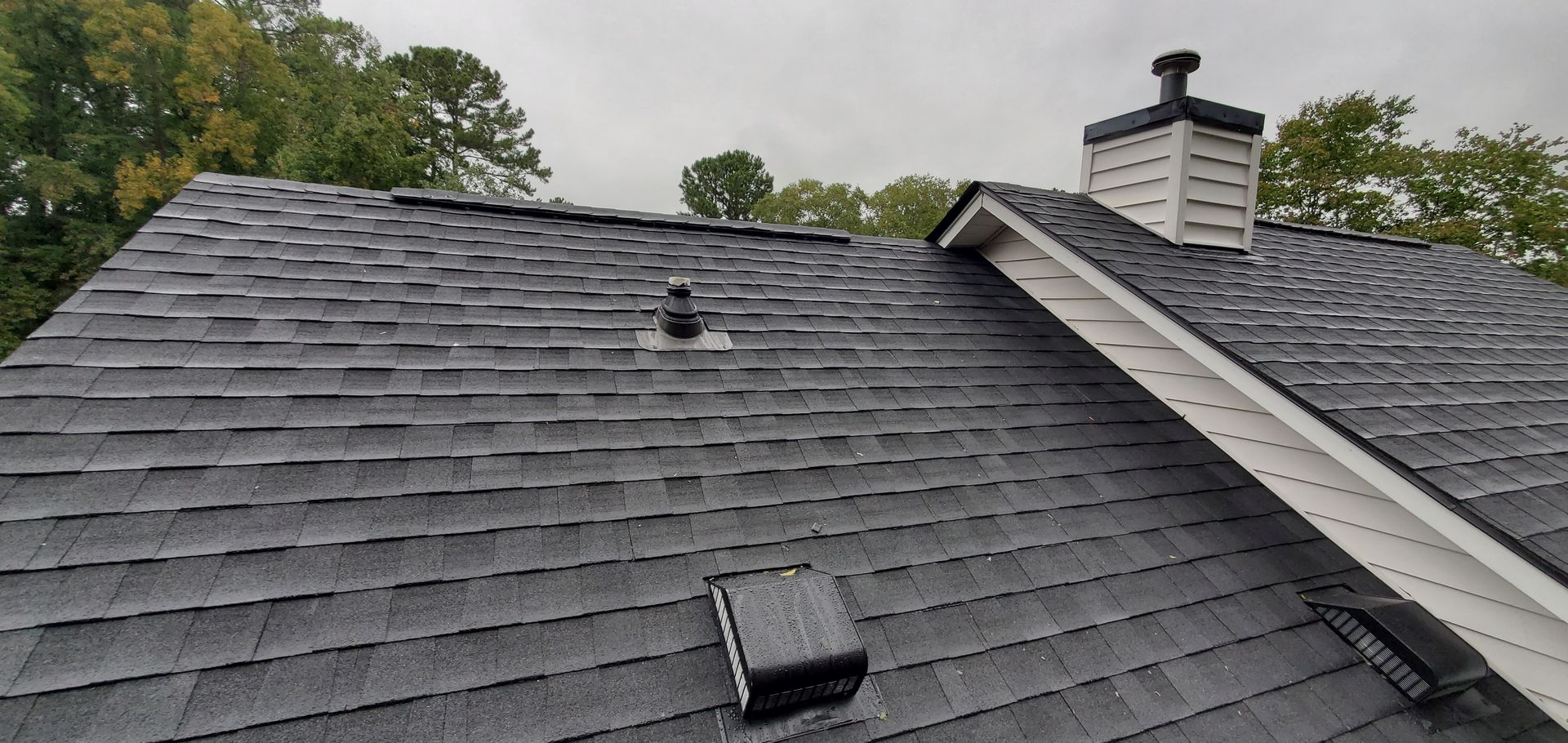 View of a dark gray shingled roof with a chimney and vent pipes under a cloudy sky.