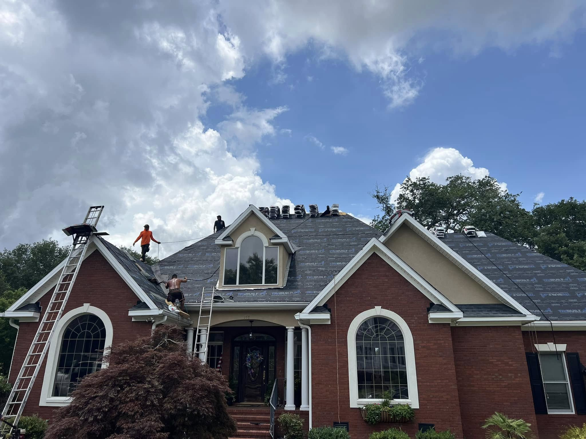 Roofers on a brick house, working on the roof under a cloudy sky.