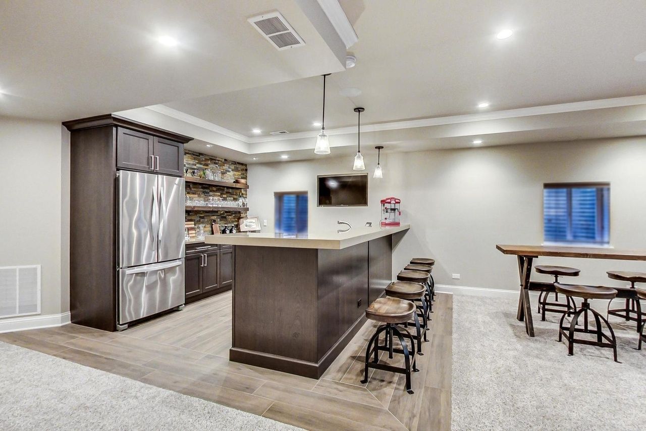 A basement with a bar , stools , a table and a refrigerator.