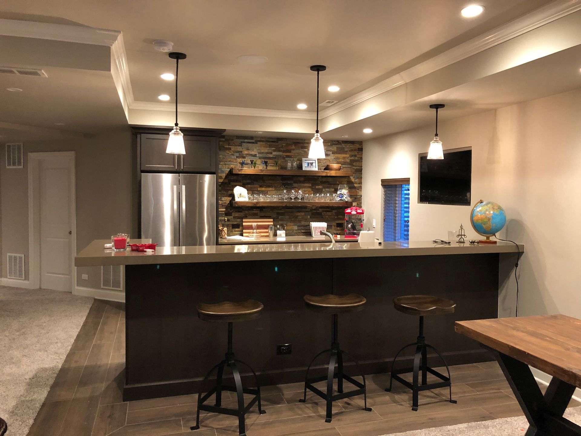 A basement with a bar , stools , a refrigerator and a table.
