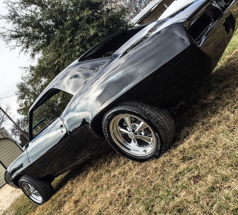 A black classic muscle car body sits on dry grass with polished chrome wheels, captured at a high angle.