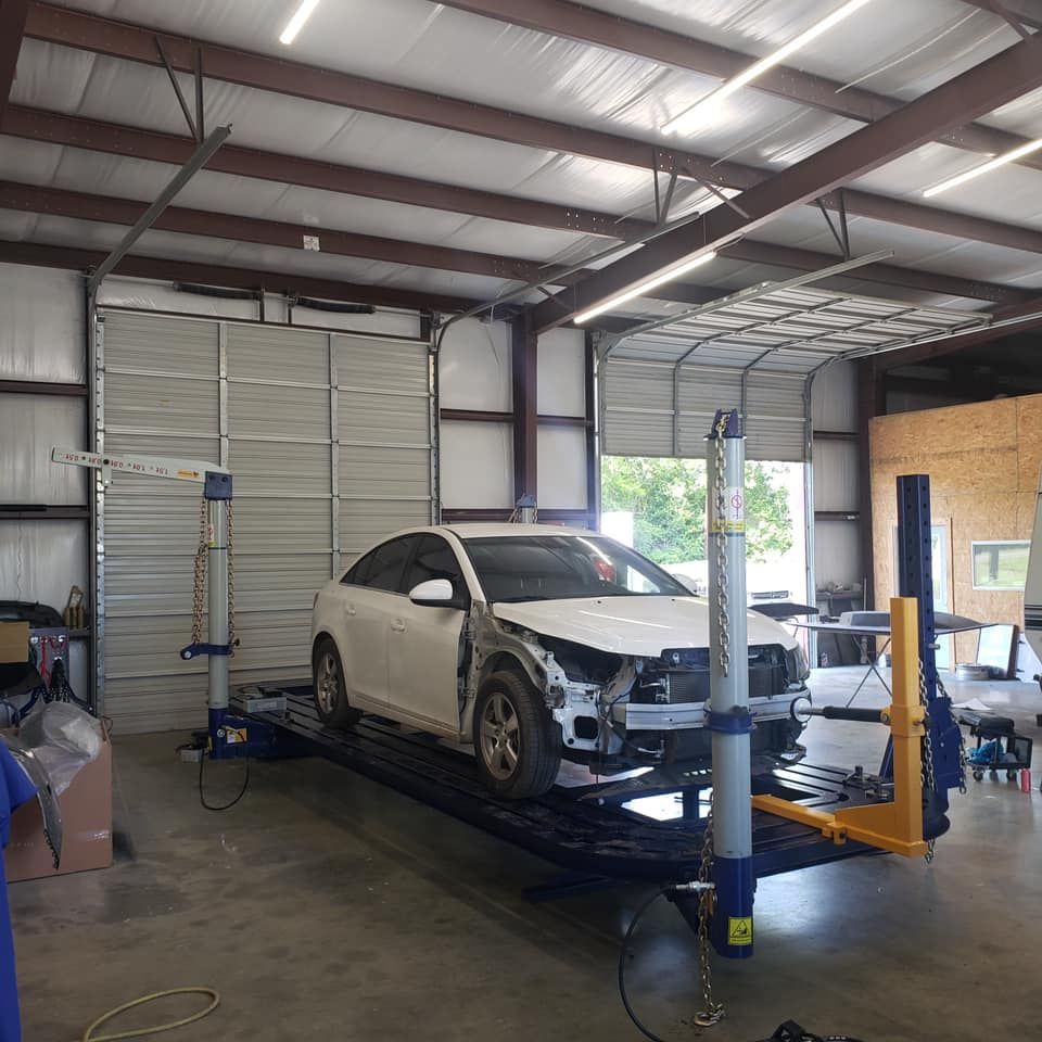 A white car with a missing front bumper sits on a metal frame alignment machine inside an auto body repair shop.