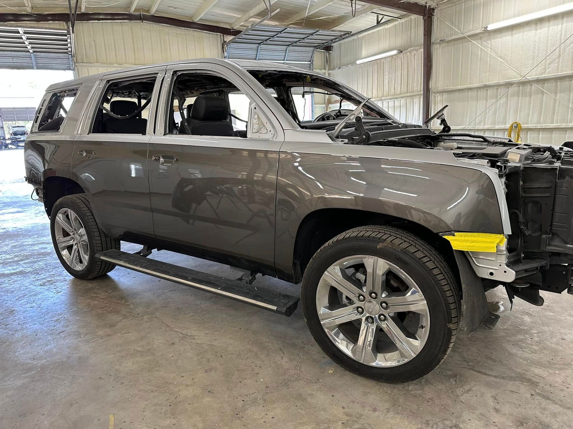 A gray SUV undergoing repairs, missing its front bumper and windows, parked inside a brightly lit auto body shop.