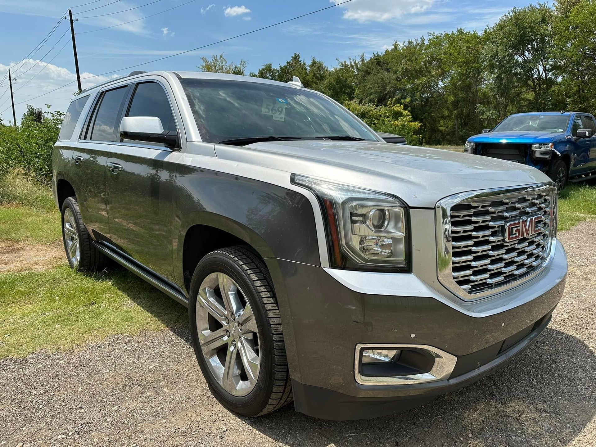 A silver GMC Yukon SUV parked on a gravel surface with trees in the background.