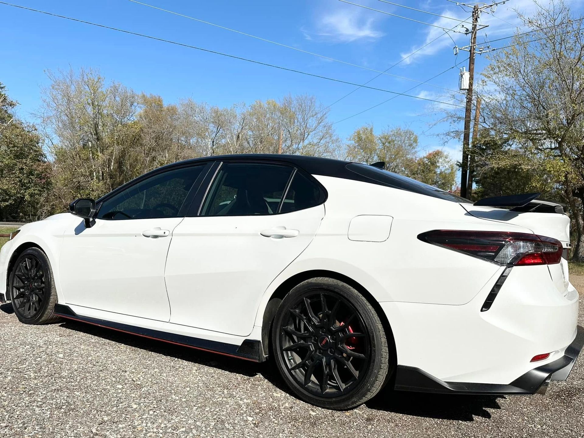 A white Toyota Camry TRD with a black roof and black wheels, parked on a gravel lot under a clear blue sky.