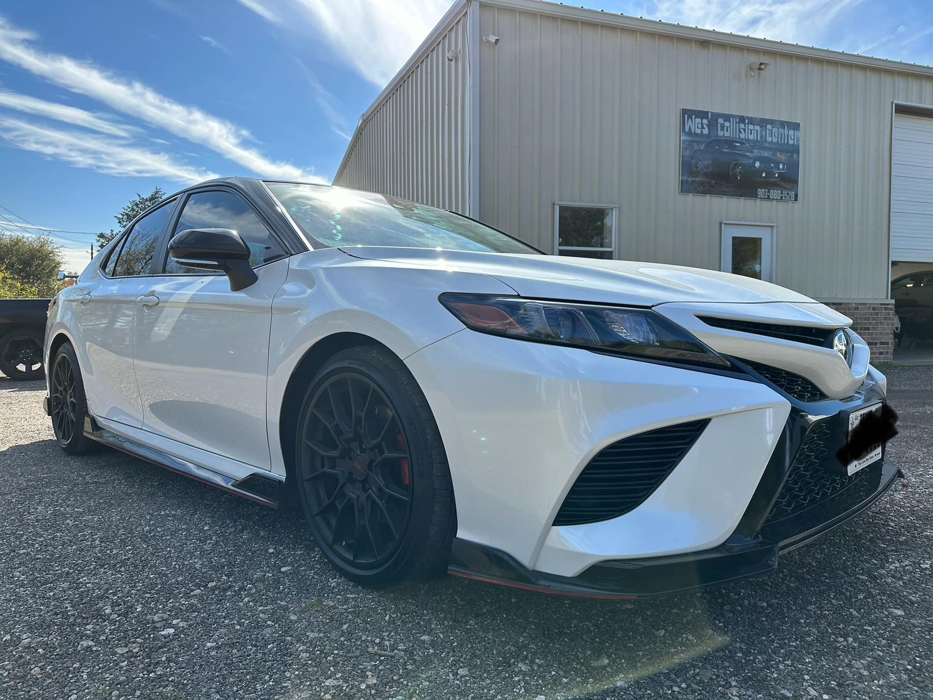 A white Toyota Camry with black wheels parked on a gravel lot in front of a metal building.