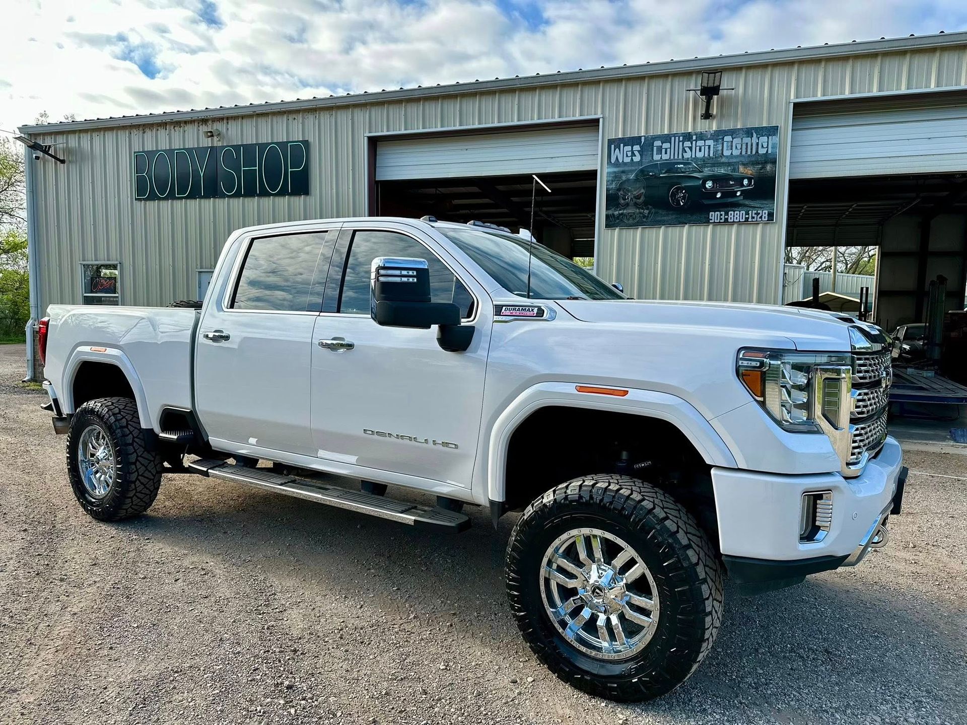 A lifted white GMC Sierra pickup truck parked on a gravel lot in front of a metal-sided automotive repair shop.