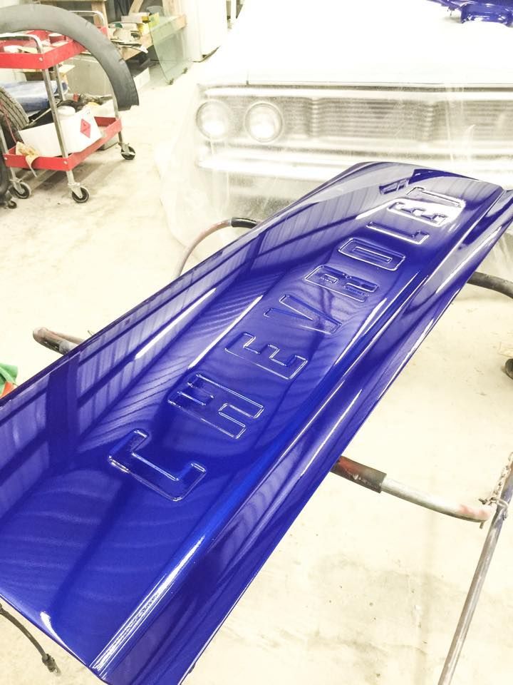 A glossy, royal blue Chevrolet truck tailgate rests on a stand in a workshop, with a car in the background.