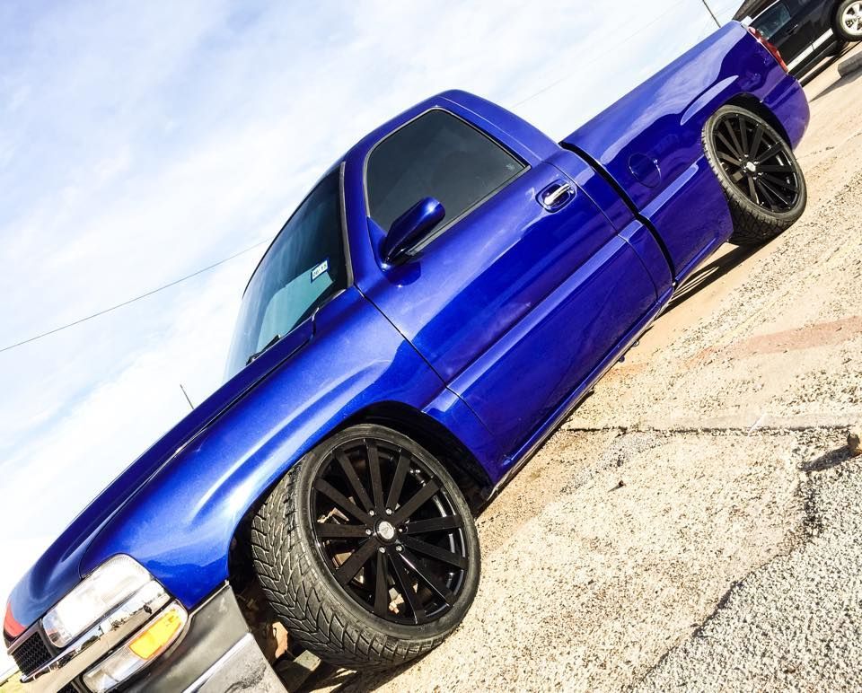 A bright blue lowered pickup truck with large black custom wheels parked on an asphalt lot.