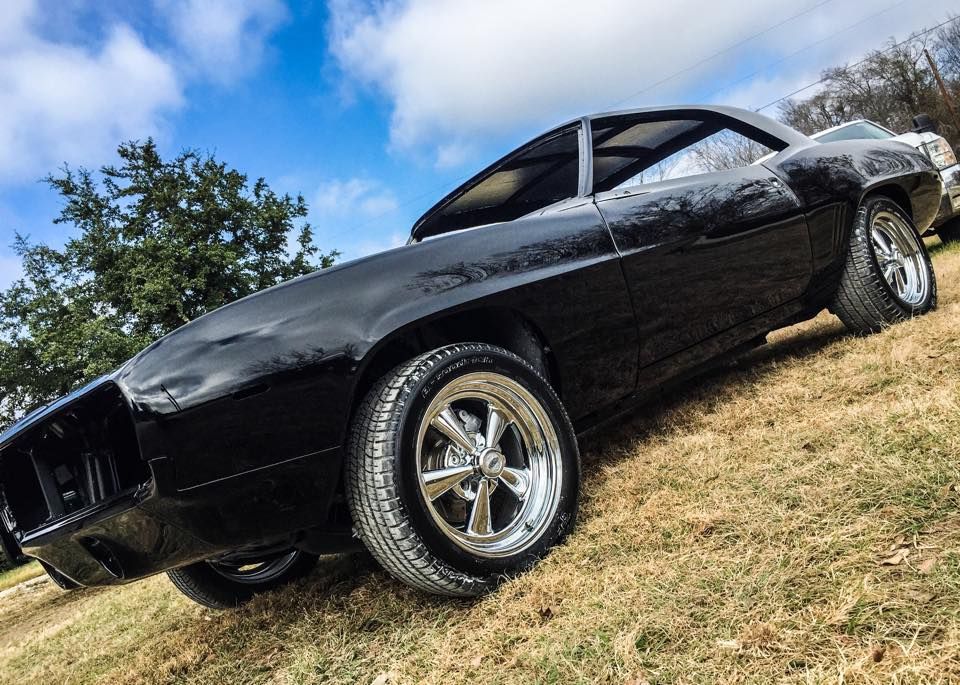 A shiny black classic muscle car parked on a grassy field under a cloudy blue sky.