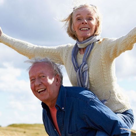 Elderly man giving woman a piggyback ride outdoors; they are smiling and appear joyful.