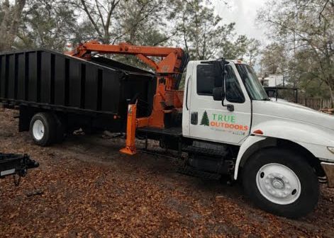 White truck with orange crane and black container in a wooded area.