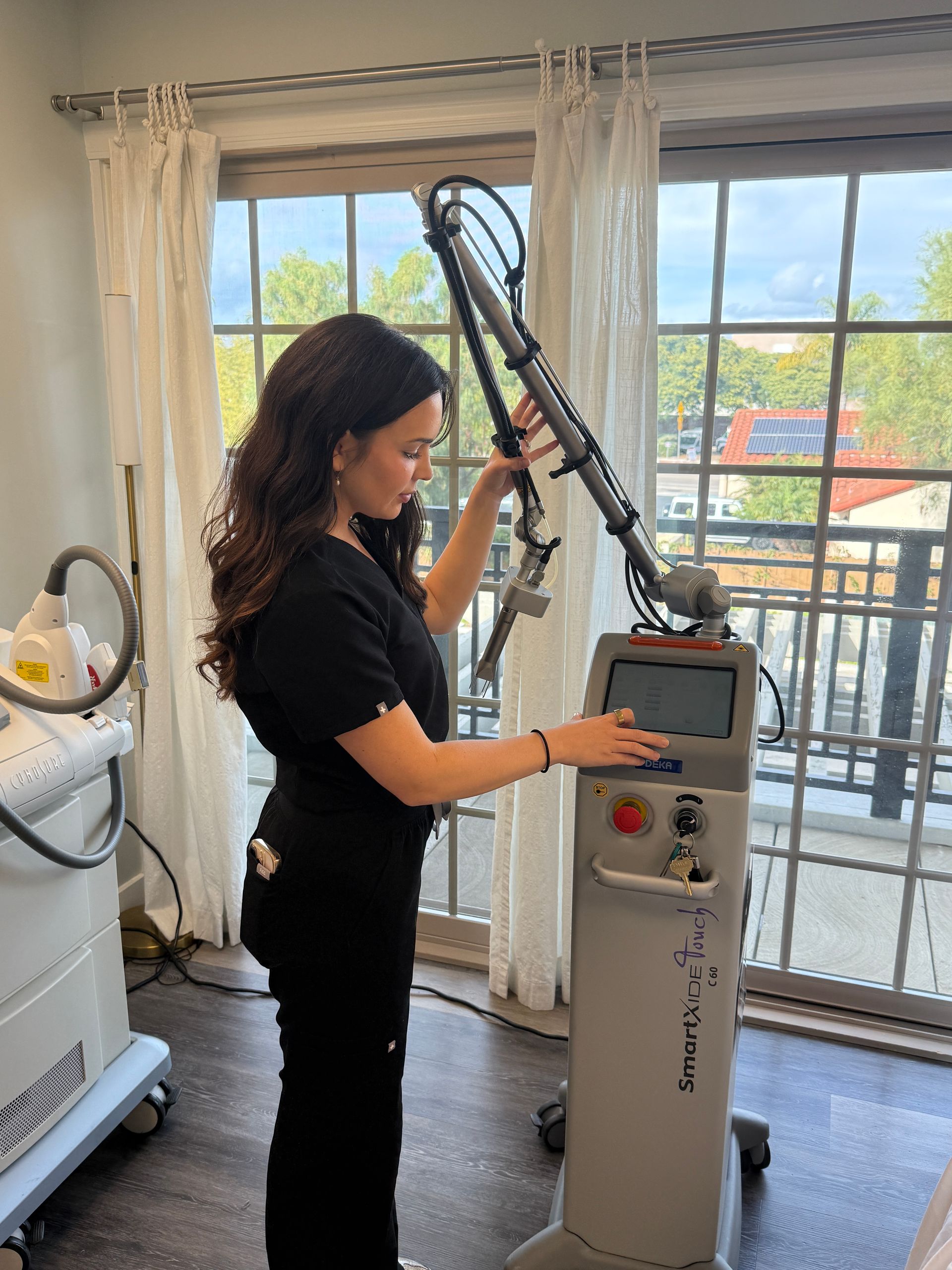 Woman in black scrubs operating a medical laser device, near a window.