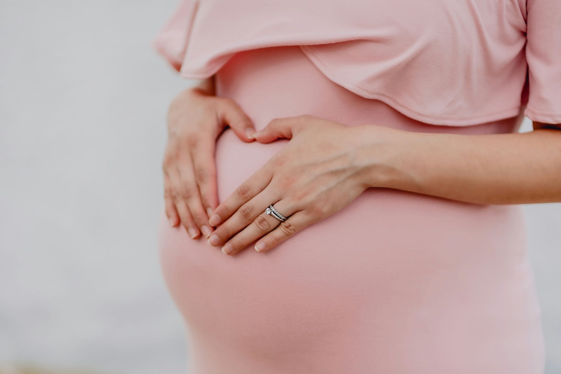 A pregnant woman in a pink dress holds her hands in a heart shape over her baby bump.
