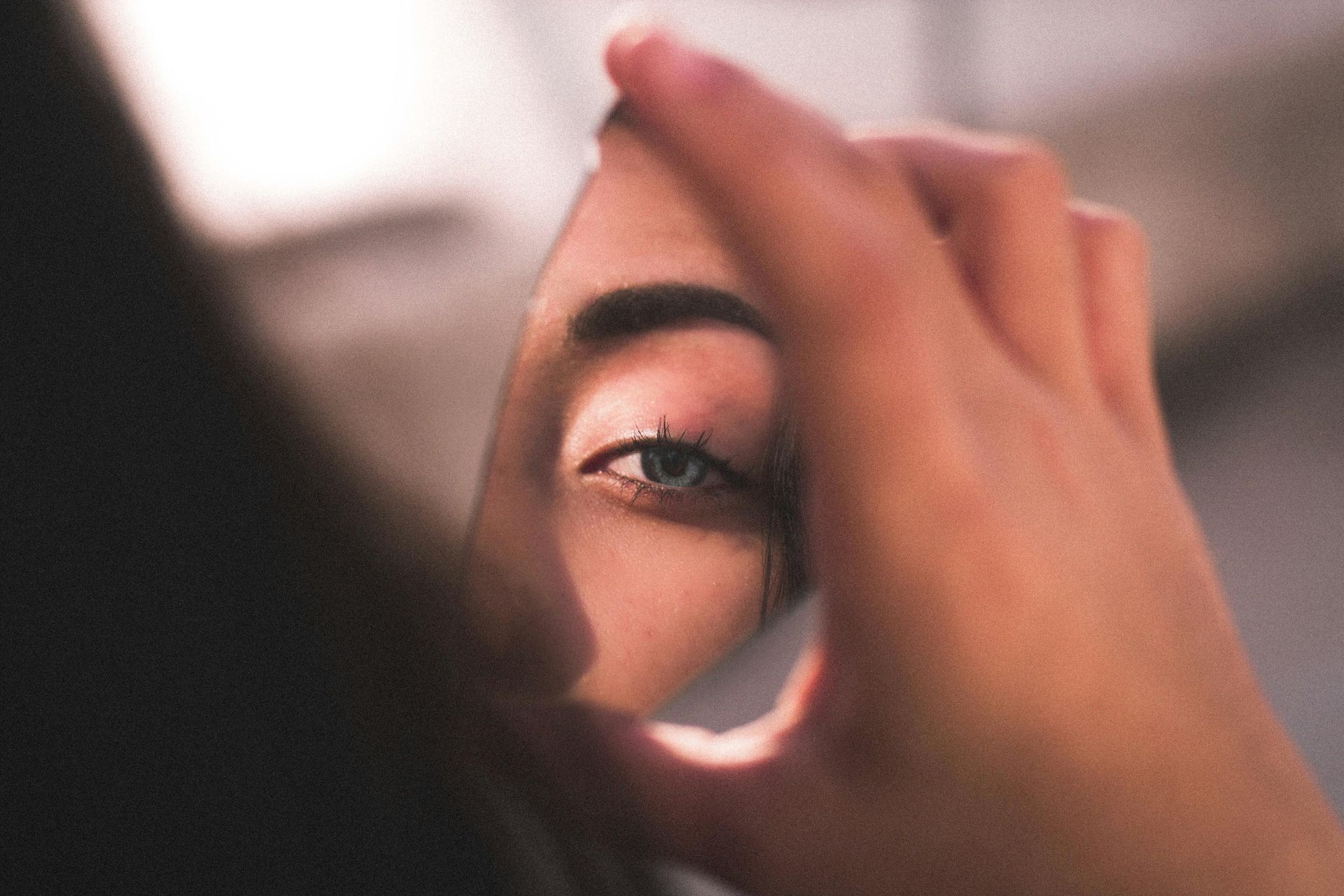 A person holding a small mirror up to their eye, blue eye visible in the mirror.