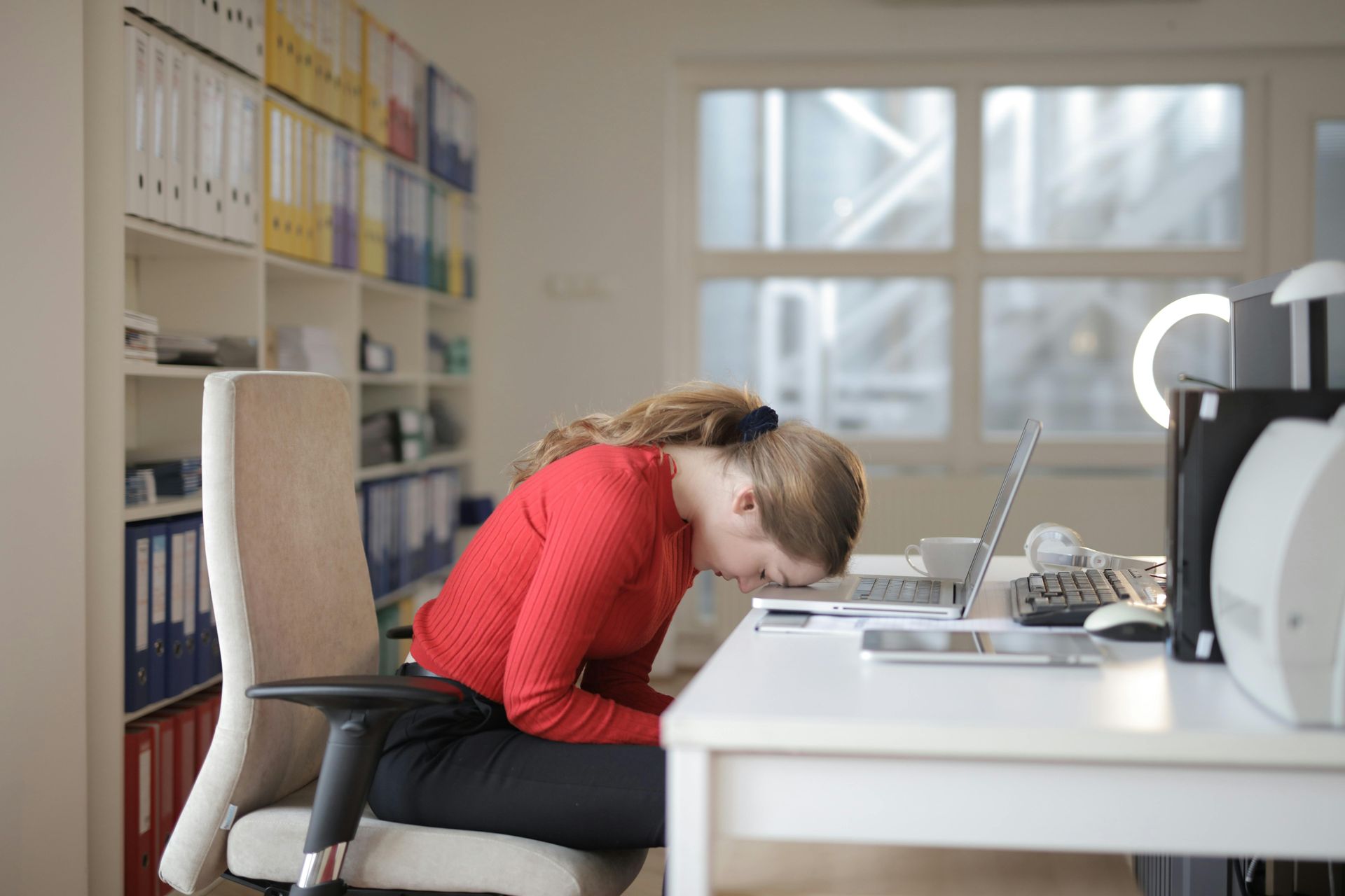 Woman in red shirt slumped at desk in an office, head down beside a computer.
