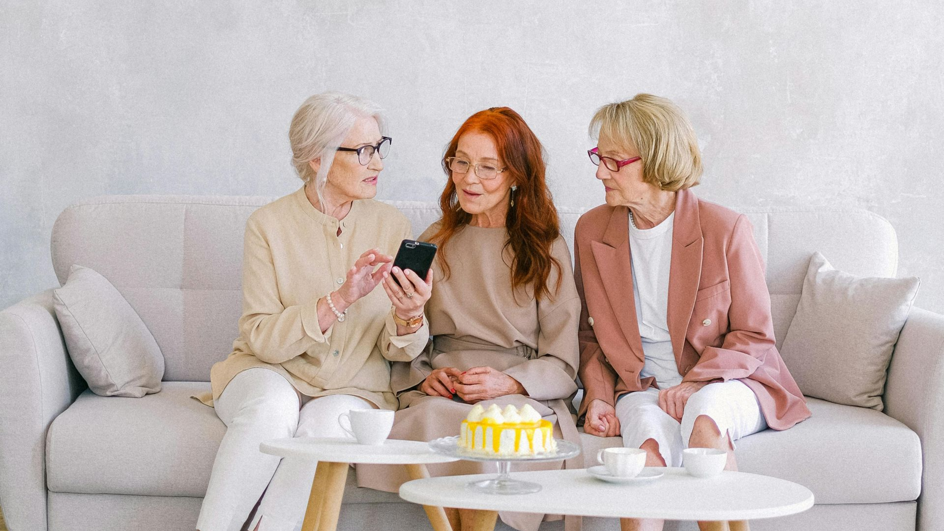 Three women look at a phone on a couch, near a cake and coffee.