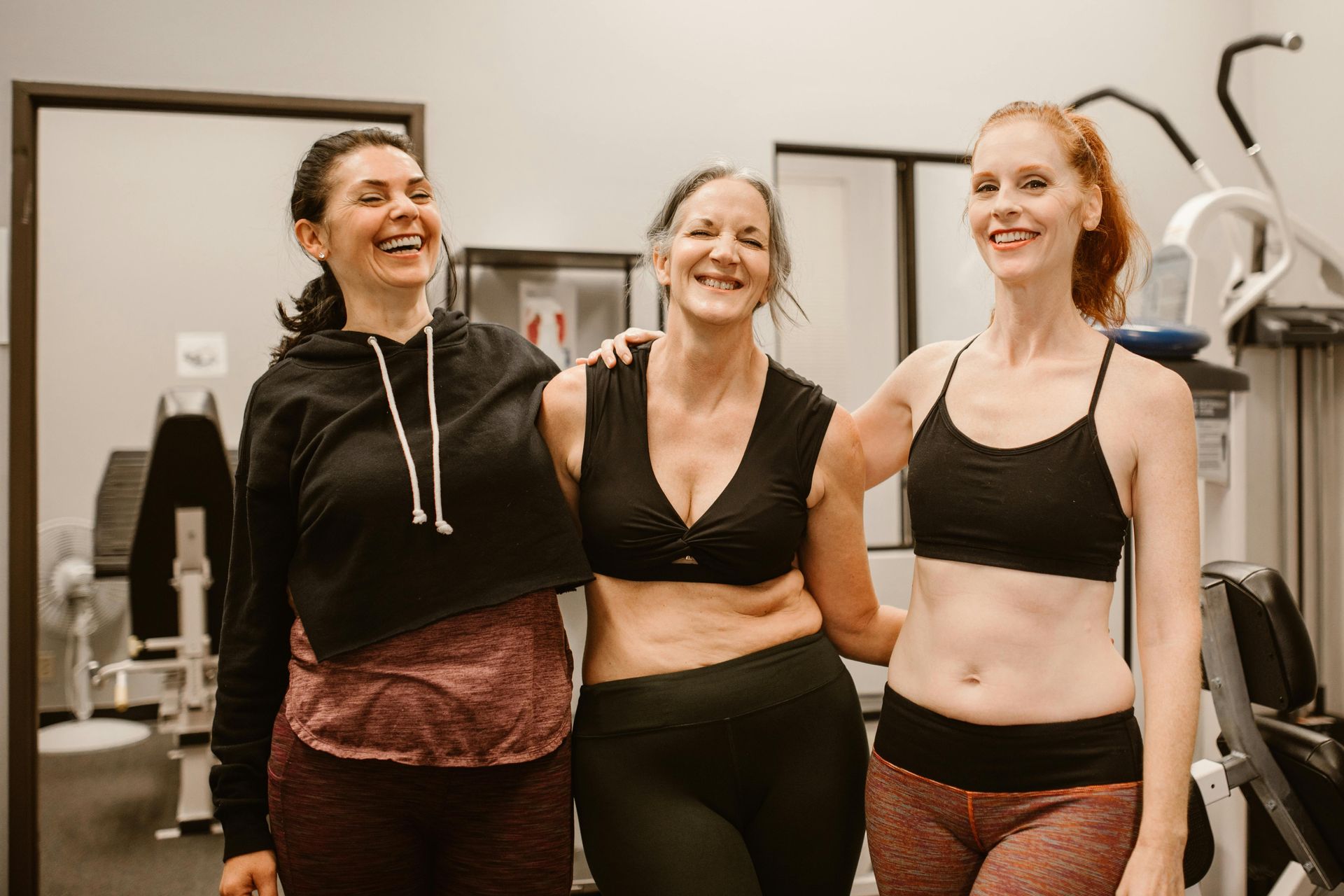 Three people in activewear stand together in a gym, smiling and resting their arms on one another.