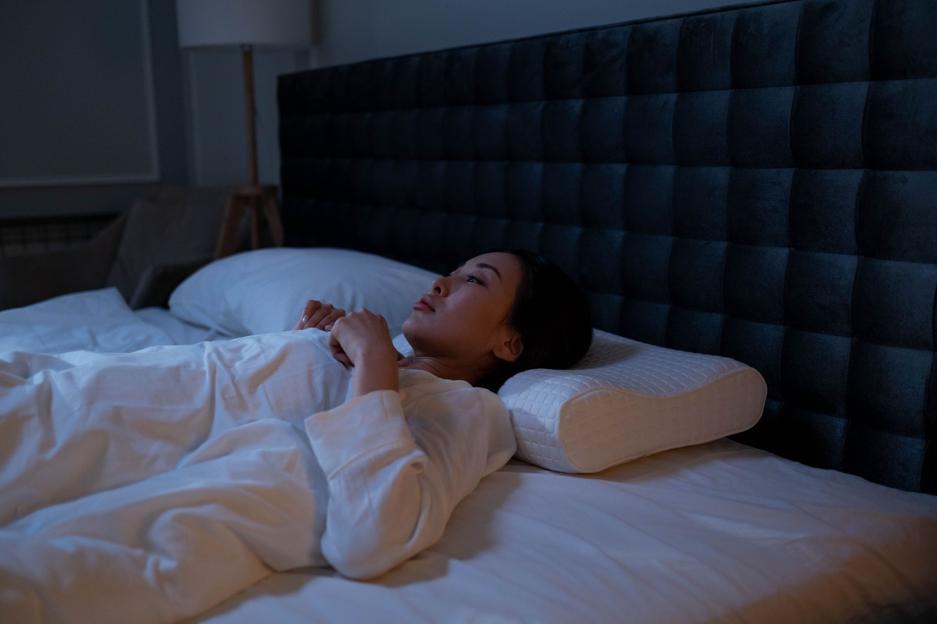 Person lying in bed under white sheets beside a gray headboard, resting in dim blue light