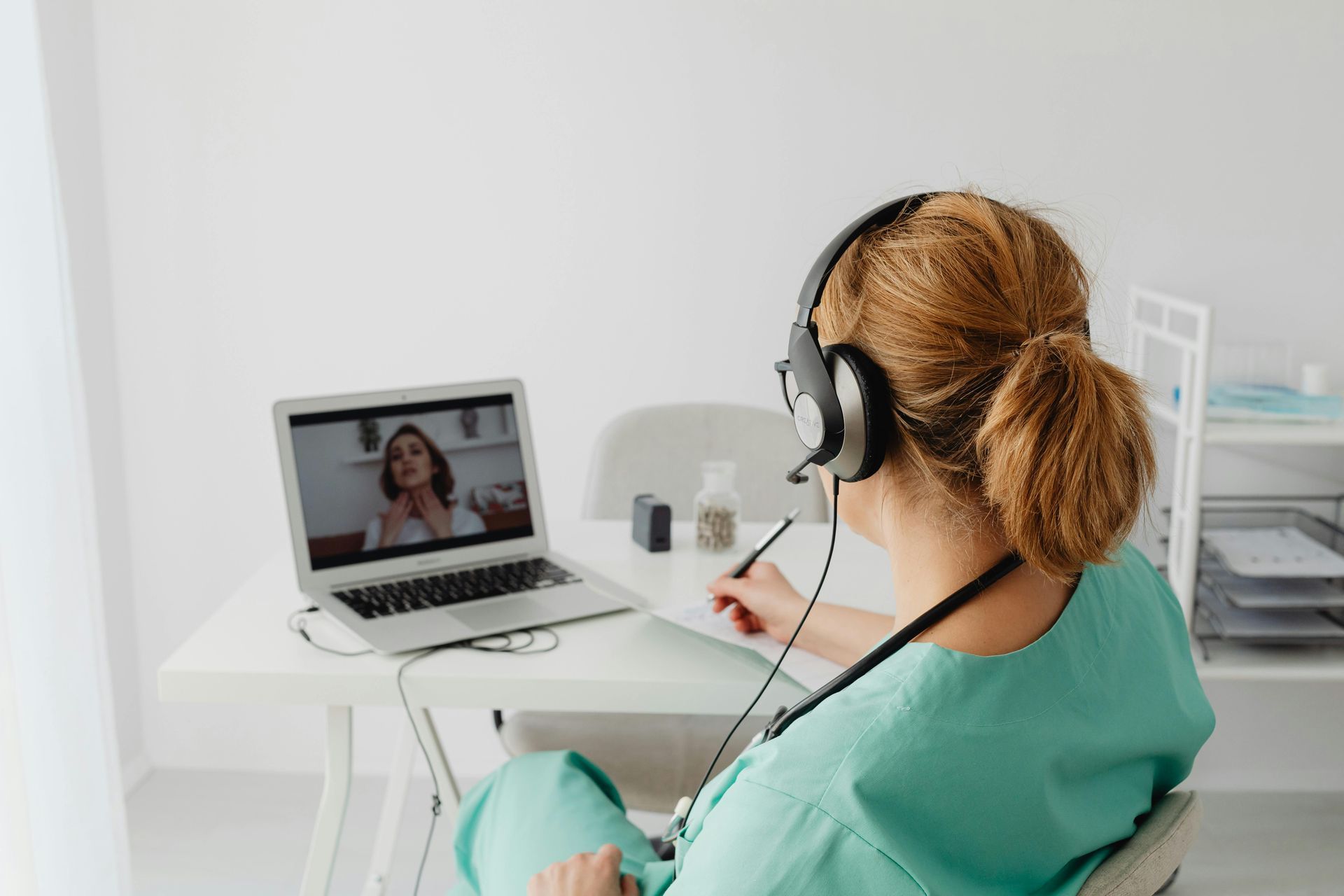 Person in green scrubs with headphones doing a video consultation on a laptop in a bright room