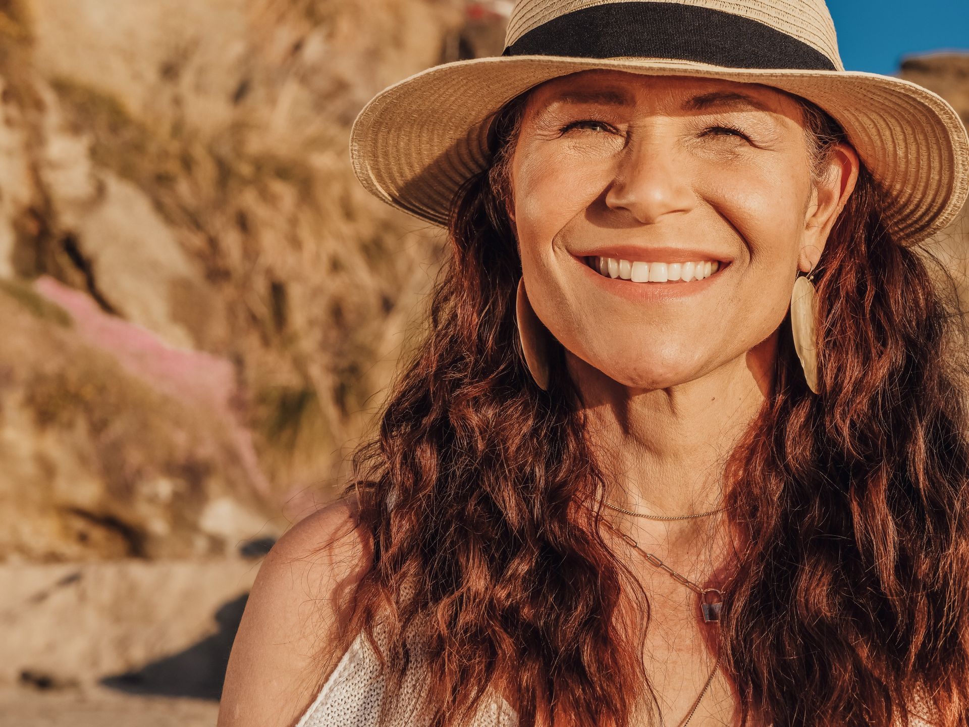 Smiling woman in a straw hat standing by rocky cliffs in warm sunlight