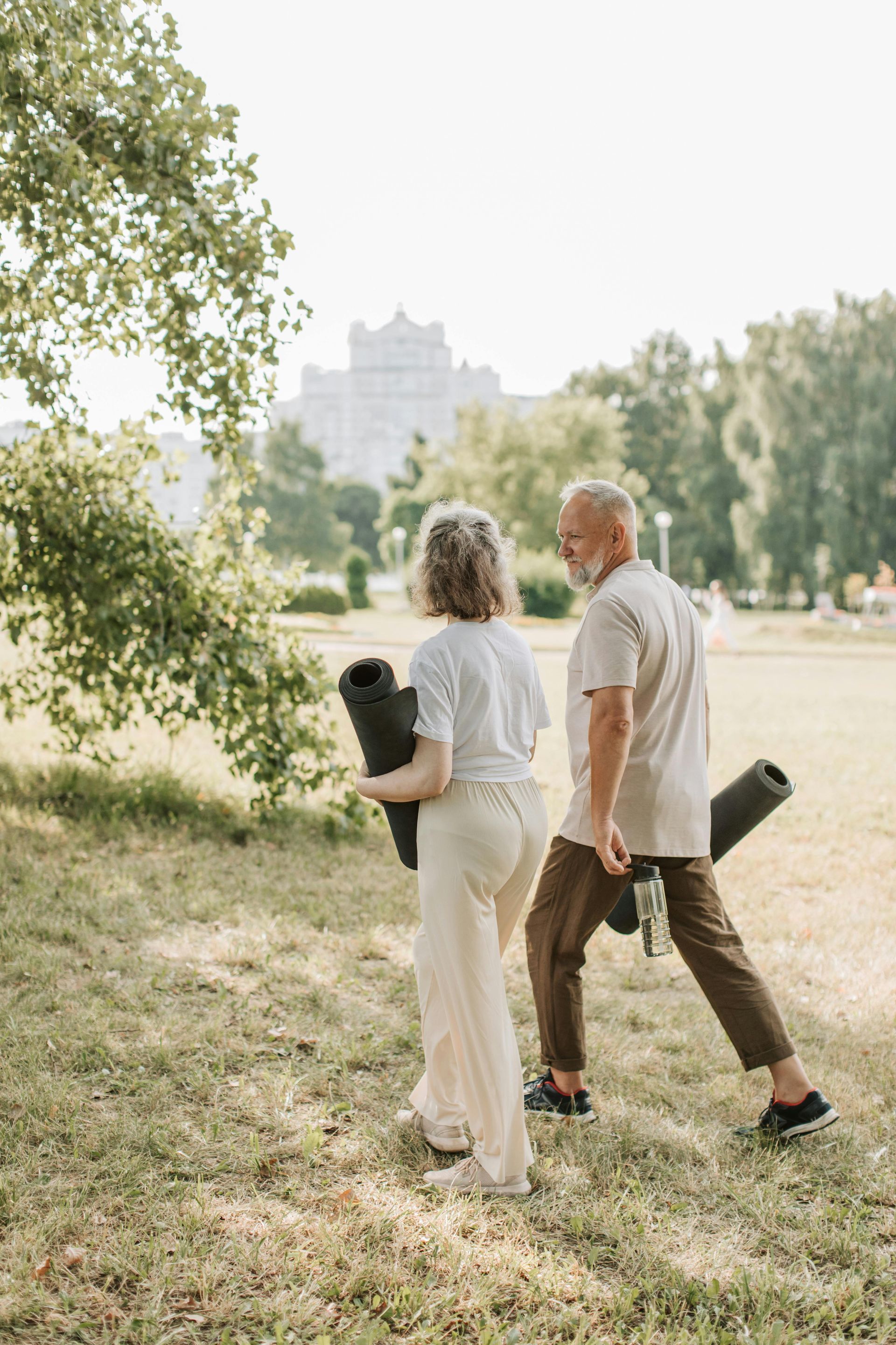 Two people walk through a park carrying yoga mats, wearing casual summer clothing.