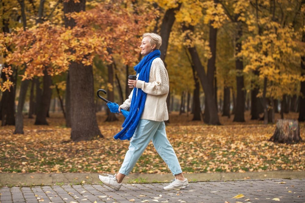 A happy middle-aged woman walking through a park while holding a coffee.