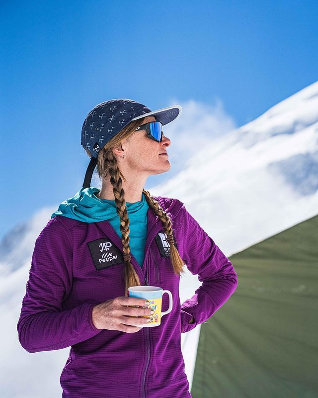 Alessandra Pepper in a purple jacket is holding a cup of coffee in front of a snowy mountain.