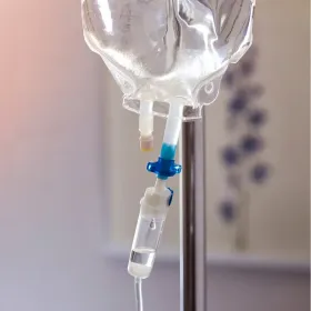 Close-up of a clear IV fluid bag attached to a drip chamber, suspended on a medical stand in a clinical setting.