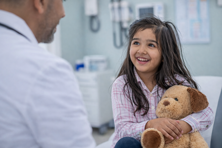 A smiling child holds a tan plush dog while sitting in a medical office and looking at a person in a white coat.
