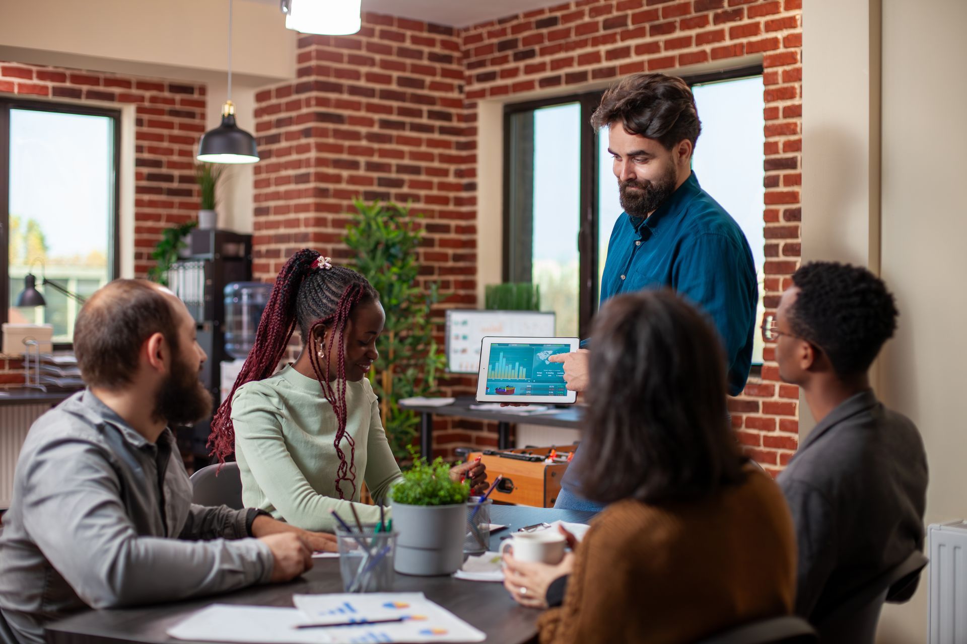 A diverse team discusses a digital presentation in a bright, modern office with exposed brick walls.