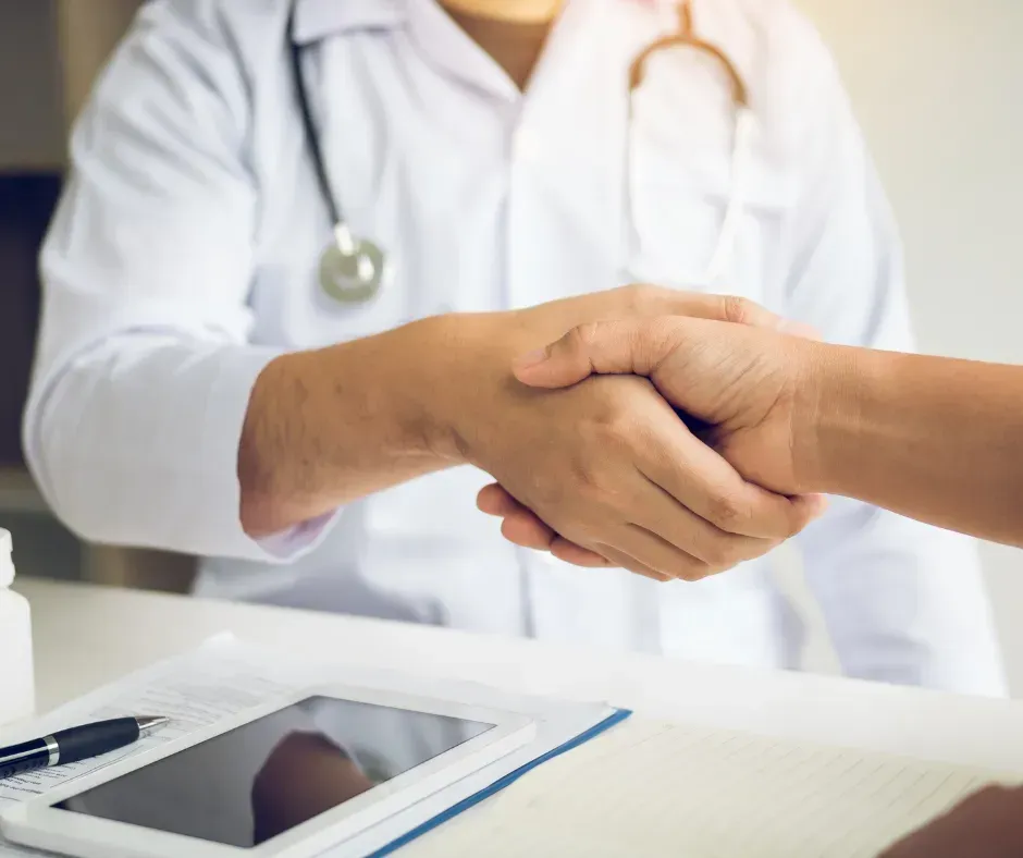 A doctor wearing a white coat and stethoscope shakes hands with a patient over a desk with a tablet and pen.