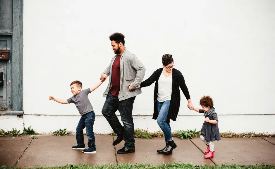 A family of four walking hand-in-hand along a sidewalk next to a white wall.