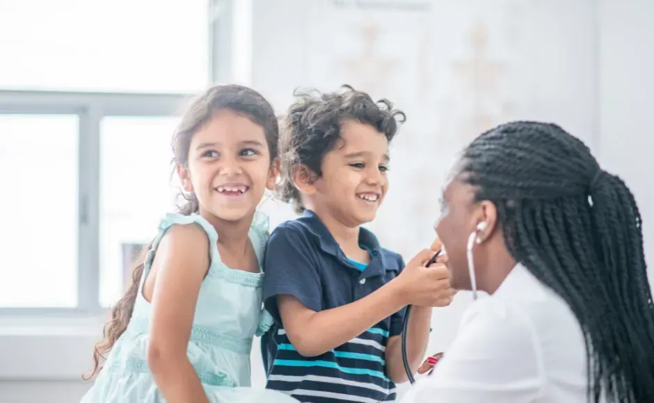 A smiling doctor examines two children with a stethoscope in a bright, modern office.