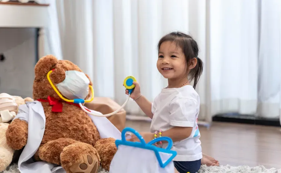 A smiling child in a white shirt holds a stethoscope to a teddy bear wearing a face mask and a white lab coat.