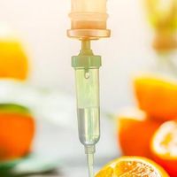 An IV drip chamber with clear fluid, set against a blurred, bright background of orange citrus fruit slices.