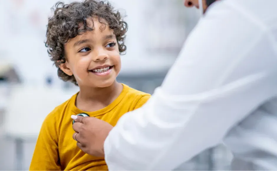A person in a white lab coat uses a stethoscope to examine a smiling child wearing a yellow shirt.