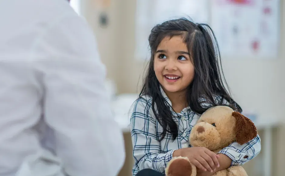A smiling child holds a tan plush dog while sitting in a medical office and looking at a person in a white coat.