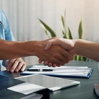 A person in blue scrubs shakes hands with another person across a desk with documents and a tablet.