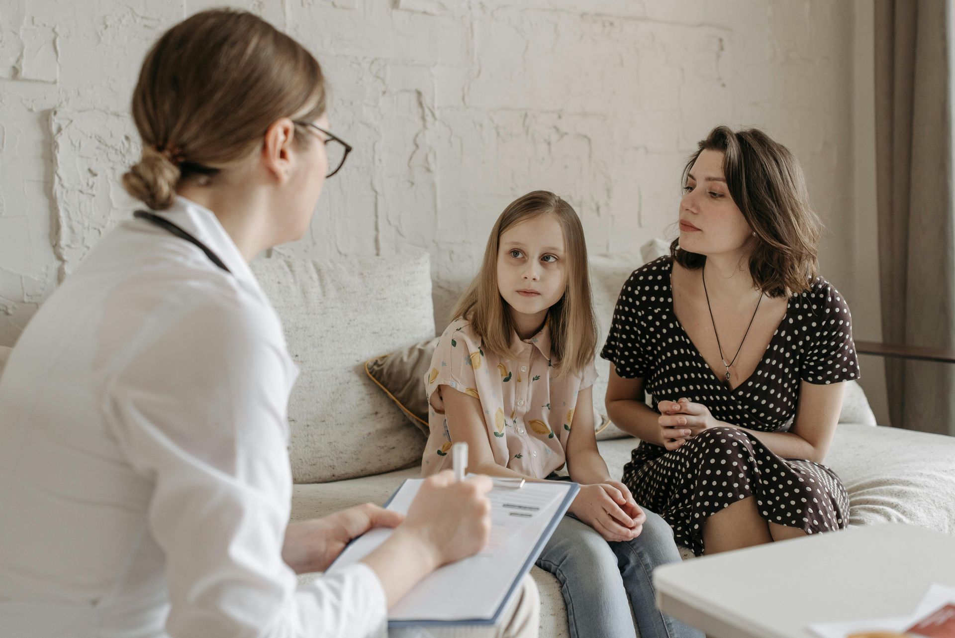A medical professional takes notes while talking to an adult and a child sitting together on a sofa.
