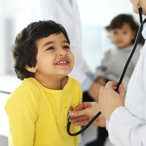 A child in a yellow shirt smiling as a healthcare professional listens to their heart with a stethoscope.
