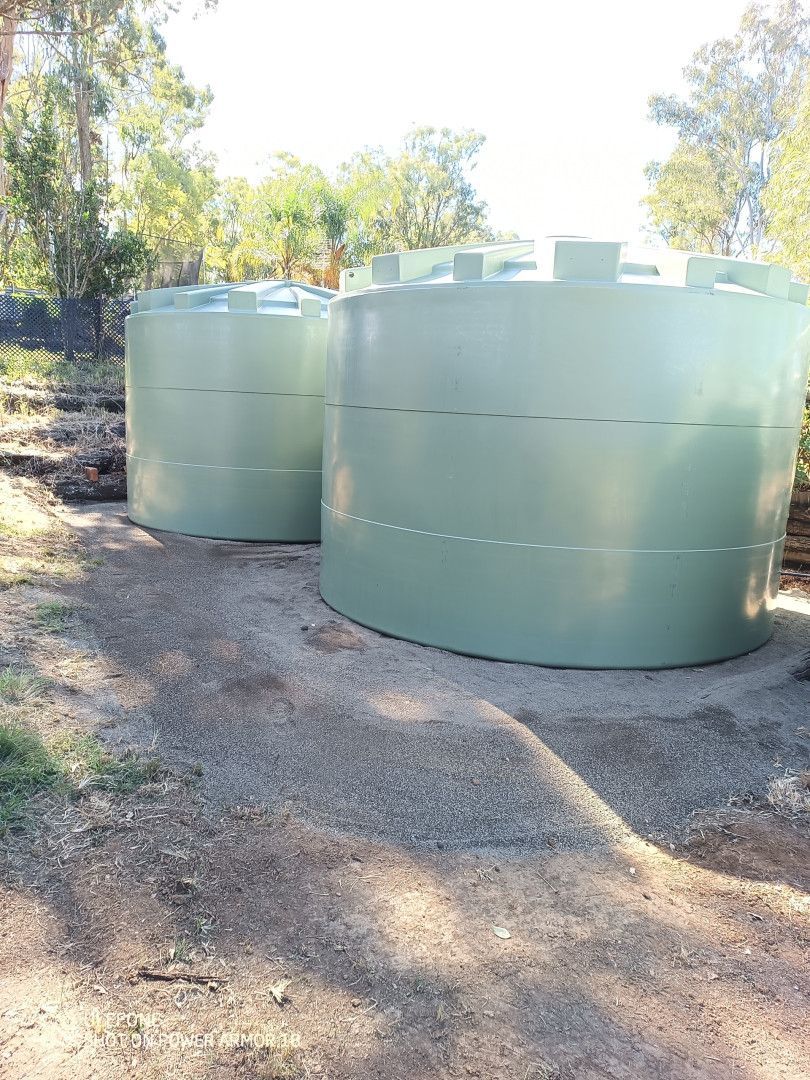 Two large, cylindrical, light green water tanks outdoors on a gravel base, trees in background.