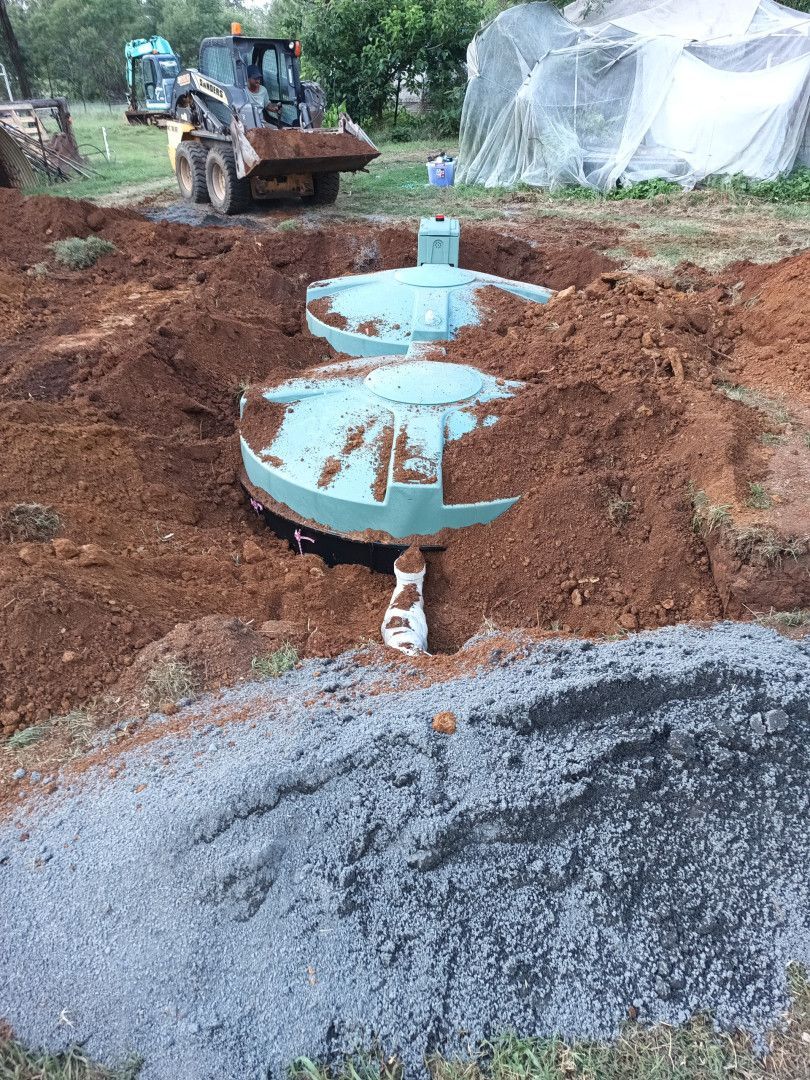 Two green septic tanks buried in soil, with a backhoe in the background.