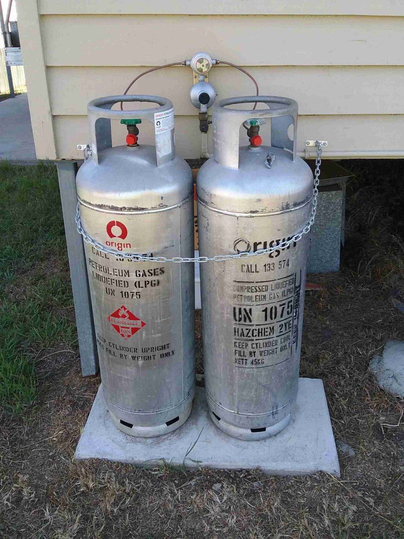 Two silver propane tanks, connected by a hose and regulator, stand on a concrete pad near a building's exterior.