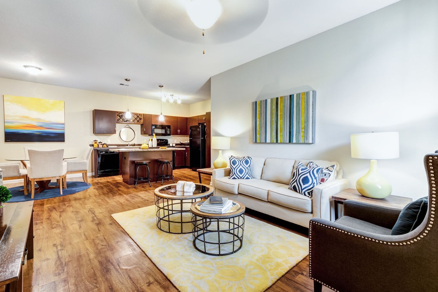 Open-concept living space with kitchen, dining area, and living room at The Preserve at Essex Farms, offers apartments for rent in Charleston, SC. Wood floors, neutral walls, yellow rug.