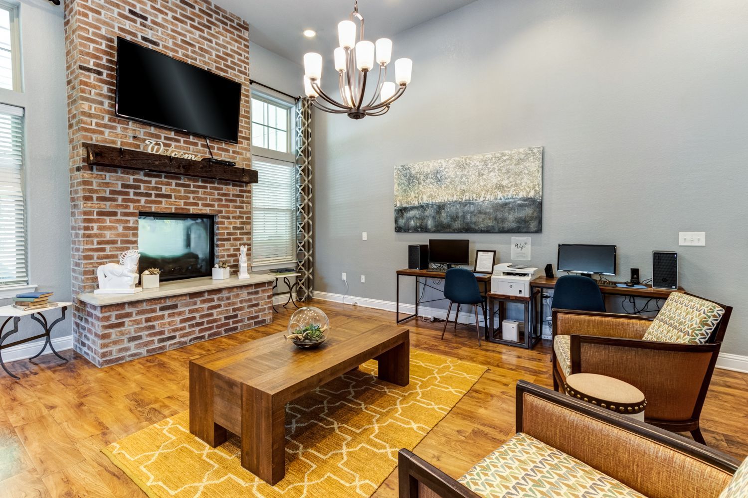 Clubhouse with brick fireplace, TV, and two armchairs, plus a workspace and chandelier at The Preserve at Essex Farms, offers apartments in Charleston, SC.